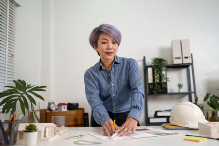 Adult Asian women architect is carefully design blueprint drawing plan construction building project with professional skill for architecture designs and construction projects. Successful women architect works hard for perfect designs.の写真素材
