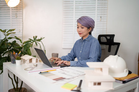 Adult Asian woman architect working on a house project on her laptop. This architect is using...
