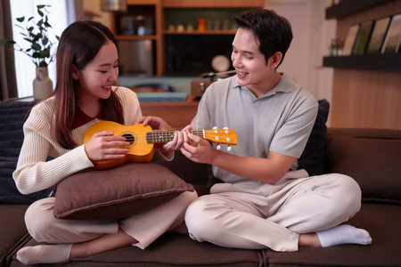 A young Asian couple is sitting on a sofa at home. The woman is holding a ukulele, and the man is showing her how to play it. They are both smiling and appear happy, enjoying their time.の写真素材