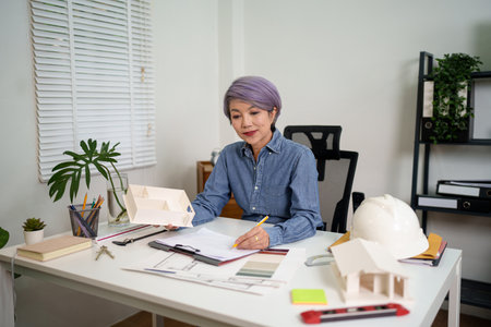 An Asian woman architect in her office designs blueprints, holding a home model. The Asian woman is focused, planning house architecture with precision. This showcases the architect's skill in construction design.の写真素材