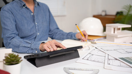An adult Asian architect using a tablet and pencil to review house blueprint design. The Asian...