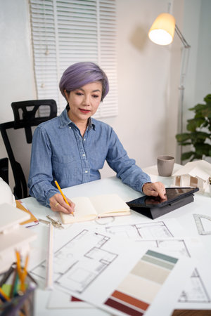 An adult Asian women architect is designing home blueprint plans and using a tablet. The Asian women architect is carefully reviewing construction documents for the project.の写真素材