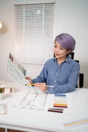 Older Asian woman architect reviewing blueprint for house design project with color palette for interior design plan. Create your best architect blueprint here with woman architect.の写真素材