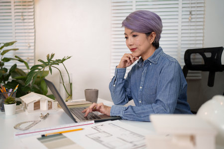 Adult Asian woman architect designs blueprints online for construction. The Asian woman interior designer creates construction plans and reviews architectural designs with a laptop on her desk.の写真素材