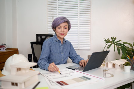 An Asian woman architect designs house blueprints with a laptop and pencil at a desk in her home office. The architect designs house building architecture floor plan designs.の写真素材