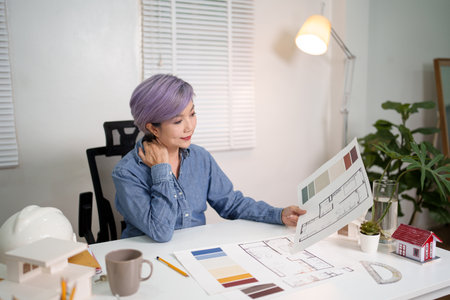 An Asian woman architect is reviewing a design blueprint for a home renovation project. The older...