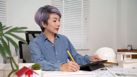An adult Asian woman architect is working on home blueprint plans at her desk using a tablet and pencil. Home design concepts and home design ideas are displayed alongside a model house. This scene shows a professional architect engaged in the design process.の写真素材