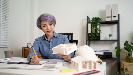 Older Asian architect woman reviewing house model blueprint plan in office. Examining construction project details for planning architecture design. She excels at architecture design, ensuring the project's success 170の写真素材