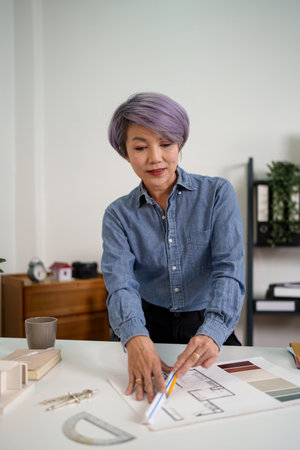 An adult Asian woman architect is diligently designing a house plans blueprint at her office table. She focuses expertly on the project. The house plans design blueprint showcases her adult skills.の写真素材
