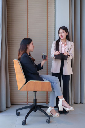 Two adult Asian women are having a business discussion in a modern office. One is sitting and holding a coffee cup, while the other is standing with a clipboard, sharing ideas and planning. Teamwork in action.の写真素材