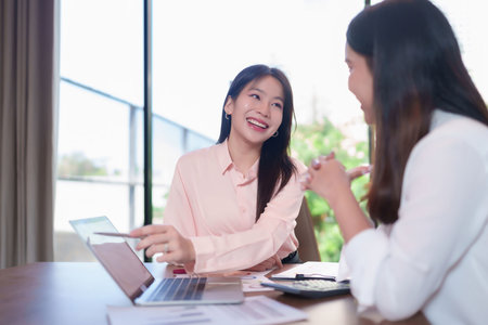 Two young Asian women are in a business meeting discussing strategy, pointing at a laptop screen. Reviewing data, brainstorming new ideas.の写真素材