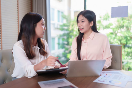 Two adult Asian women in a business meeting discussing strategy. They are collaborating on a project, brainstorming ideas with a laptop and presentation charts in a modern office.の写真素材