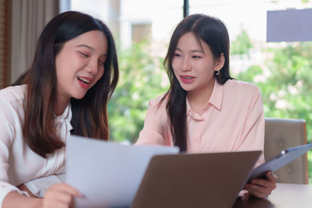 Two young Asian women are in a business meeting, analyzing data and discussing an investment plan. They're collaborating at the office, using a laptop and documents to review the information.の写真素材