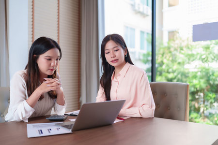 Two young Asian women in a business setting analyzing data on a laptop. They are discussing financial strategies and reviewing charts graphs for growth.の写真素材