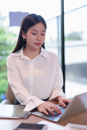 An image featuring a young Asian woman attentively working on her laptop in a modern office. Focused and dedicated, She is a businesswoman or entrepreneur working with technology.の写真素材