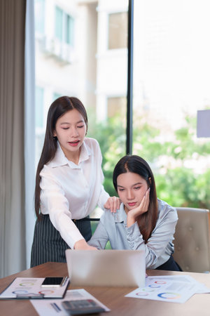 A photo shows Asian adult women in a business setting discussing problems and analyzing financial reports. The team works on a laptop with charts on the desk, showcasing workplace collaboration and support.の写真素材