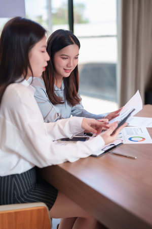 Two young Asian women in business attire reviewing charts and graphs at a desk. Analyzing data, discussing strategy. Office setting showcasing teamwork and collaboration for success.の写真素材