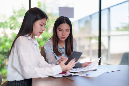 Two young Asian women analyzing business data with graphs, charts, and calculator. Discussing strategy, teamwork, and financial planning in modern office. Data analysis and success.の写真素材