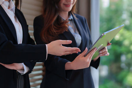 Two young Asian businesswomen in suits discuss business using a tablet. Focus on teamwork, corporate strategy, and planning. Professional communication and solutions.の写真素材