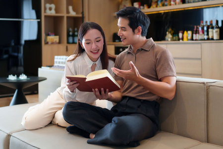 An Asian adult couple is spending quality time together reading a book on the couch at home. They appear relaxed and happy enjoying a quiet moment indoors readingの写真素材
