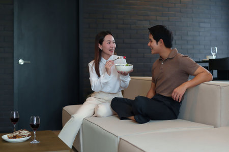 An Asian young couple is sitting on a comfortable sofa in a modern living room. They are sharing a salad and enjoying a glass of red wine creating a happy and relaxed atmosphere.の写真素材