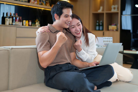 A young Asian couple sitting on a sofa celebrating success on a laptop at home. They are smiling and...
