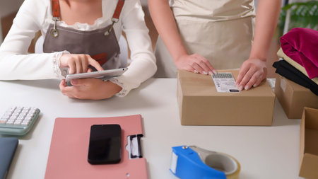 Asian adult women using a tablet to manage orders, check stocks and preparing boxes for delivery to...