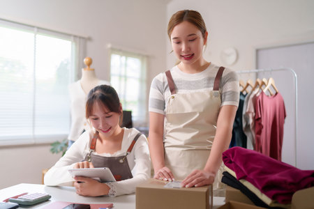 Two young Asian women are working together, managing their online fashion business. They are packing orders and using a tablet to check the stock. They ensure smooth delivery to customers with care.の写真素材