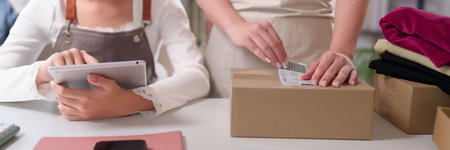Young Asian women are preparing packages for shipping. The online seller confirms orders on a tablet and affixes labels to the boxes. Home office operations are streamlined for dispatch.の写真素材