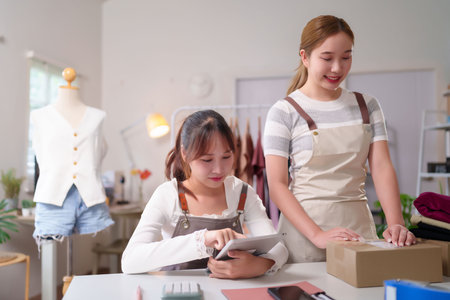 Two young Asian women are checking online orders on a tablet and preparing packages for shipping in their small business They are working with parcel boxes and other supplies for online marketingの写真素材