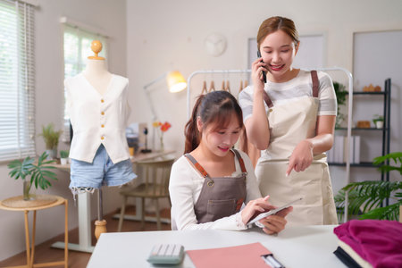 Two young Asian women entrepreneurs in their clothing design studio talk about their business on a mobile phone and tablet with mannequins and clothes nearbyの写真素材