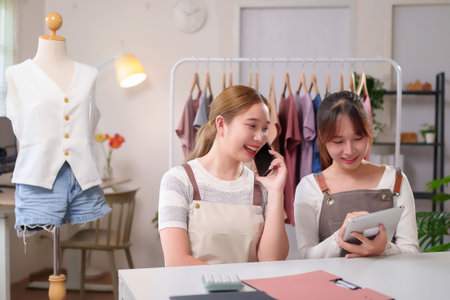 Two young Asian women are managing their online fashion store business. One is on a mobile phone, possibly taking orders, while the other uses a tablet to manage sales and stock in the studio.の写真素材