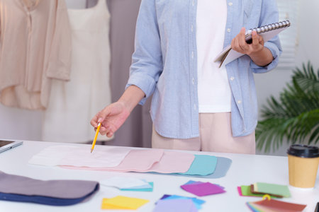 Asian adult fashion designer selects fabric samples for a clothing project. See color swatches and...