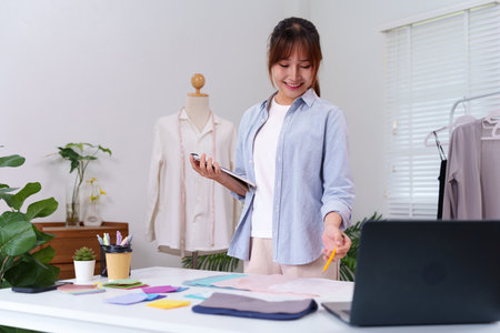 Young Asian woman is working on new clothing designs. She is surrounded by fabric swatches and other design materials. Fashion design pattern making and sewing processの写真素材