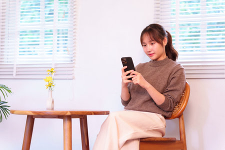 Capturing an Asian young adult woman engaged with her smartphone in a comfortable indoor environment. This image portrays modern daily life integrated with artificial intelligence for seamless communication and digital activities.の写真素材