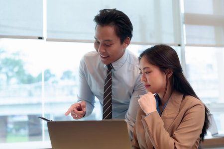 Two Asian adult colleagues working together discussing project strategy reviewing data analyzing reports on a laptop in an office environmentの写真素材