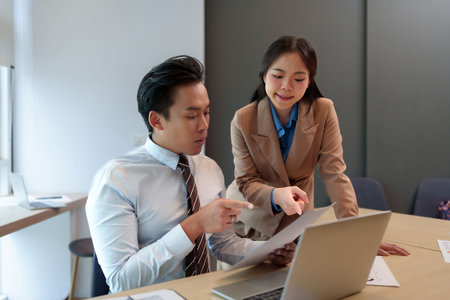 Two Asian adult professionals collaborating in office meeting discussing market data analysis reviewing business strategy planning future growth productivityの写真素材