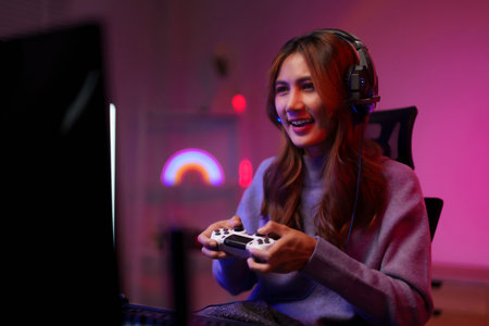 Adult Asian female streamer enjoying online esports using game console controller headset in a dimly...