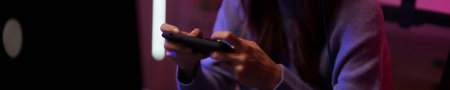 Close up view young asian hands playing mobile video game dark room vibrant neon lighting digital...