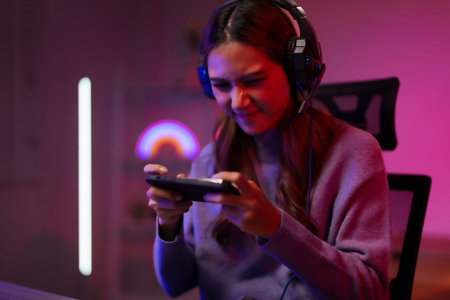 Young Asian female stream player plays mobile game online using gaming headset in a dark room with...