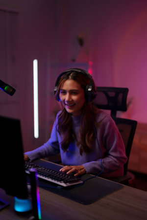 Happy Asian woman streams video game at home night using computer keyboard headset illuminated with colorful lighting setupの写真素材