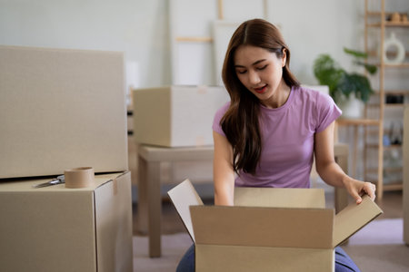 Young Asian woman carefully packing belongings into cardboard boxes for relocation preparing to move...
