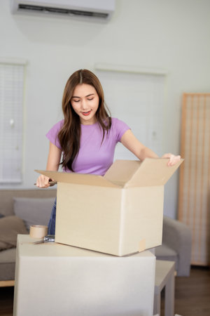 An Asian young woman packs stuff into cardboard boxes during house moving to new apartment Getting...