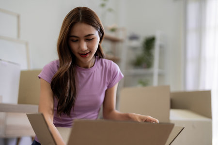 Young Asian woman unpacking cardboard boxes after moving into new home apartment house organizing...