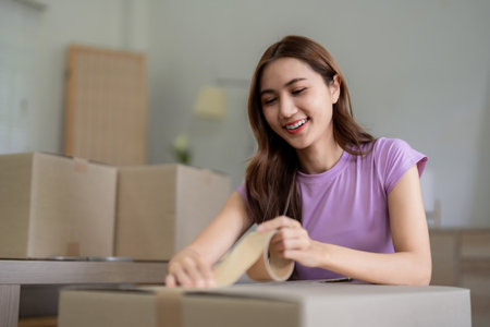 Happy Asian women preparing moving boxes for relocation to a new home sealing packages with tape...