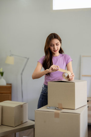 Young Asian woman diligently seals cardboard boxes with packing tape preparing for home relocation...