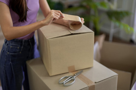 Asian woman carefully sealing cardboard box with packing tape for moving house. Preparing for new...