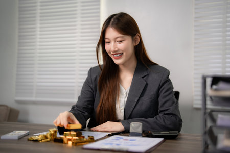Happy Asian woman financial advisor arranging gold bars coins. Business office setting demonstrates...