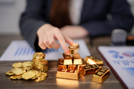 Adult hand arranges gold bars and coins on a desk with financial charts. Represents investment...