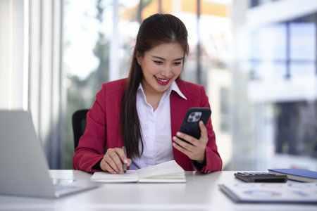 Adult Asian professional woman skillfully balances smartphone usage with note taking for efficient business operations in her contemporary office space.の写真素材